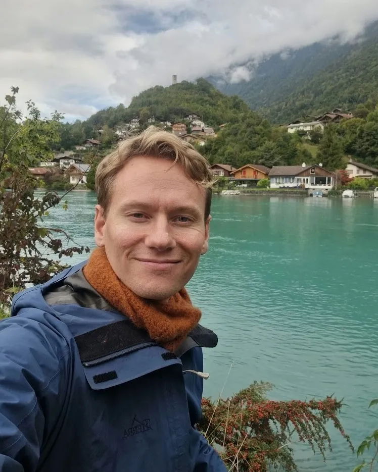 Olof by a turquoise lake, with a village and misty mountains behind him.
